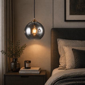 May include: A contemporary pendant light featuring a smoky grey glass globe. The light fitting has a gold-coloured metal base and an exposed Edison-style bulb. The lamp hangs above a bedside table with books and a candle, creating a warm atmosphere.