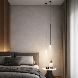 Puede incluir: Una escena de dormitorio moderno con una cama con ropa de cama y almohadas grises. Dos lámparas colgantes negras y minimalistas cuelgan del techo. Una pequeña mesa auxiliar contiene objetos decorativos. Las paredes son de tonos neutros con paneles de madera.