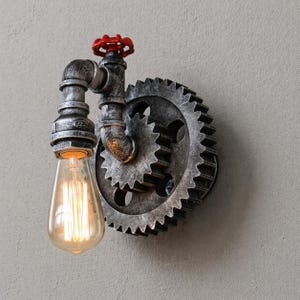 Puede incluir: Aplique de pared de estilo industrial con un engranaje grande, tuberías metálicas y una válvula roja. La lámpara tiene una bombilla Edison vintage. El metal tiene un acabado plateado envejecido.
