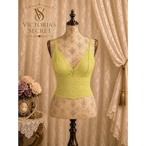 May include: A lime green lace bralette featuring a V-neckline with a small center bow detail. The garment is displayed on a mannequin and showcases an intricate floral lace pattern throughout the bodice with a scalloped hemline edge.