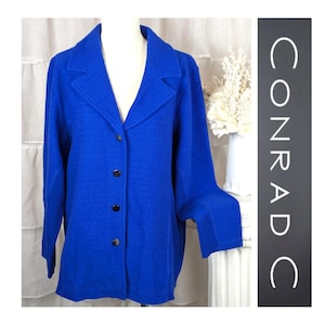 Peut inclure: Un blazer bleu vif avec un col cranté classique et trois boutons noirs. La veste à manches longues est présentée sur un mannequin, mettant en valeur sa coupe ajustée et son tissu texturé. Le mot "CONRAD C" est imprimé sur le côté droit de l'image.