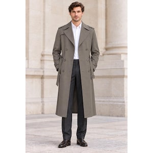 May include: A long, double-breasted trench coat in a muted olive-gray tone, featuring wide lapels, shoulder epaulets, and a belted waist. The coat is worn over a crisp white button-down shirt and dark charcoal gray trousers with dark leather shoes.