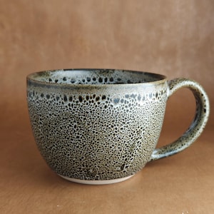 May include: A white ceramic mug with a black handle and the text "But First, Coffee" in a black, bold font. The mug is sitting on a wooden surface. The mug is approximately 4 inches tall.