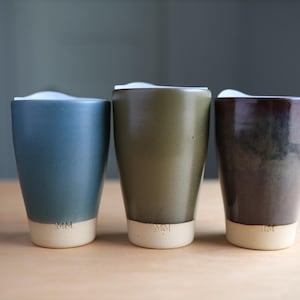 May include: Three ceramic tumblers in shades of blue, green, and brown, each with a cream-colored base. The tumblers have a smooth, matte finish and a simple, modern design. The tumblers are approximately 5 inches tall.