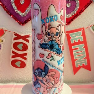 May include: Pink and white tumbler with a glitter finish featuring a cartoon couple of Stitch and Angel from the Disney movie Lilo & Stitch. The tumbler has a variety of hearts, flowers, and the words "XOXO" and "Be Mine" printed on it.