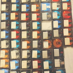 Könnte beinhalten: Eine Ansammlung von Sony MiniDisc-Hüllen, jede mit einer Disc. Die Discs haben verschiedene Farben, darunter Blau, Orange und Rot. Jede Hülle hat ein kleines Papieretikett. Die Zahl "74" ist auf einigen Hüllen aufgedruckt.