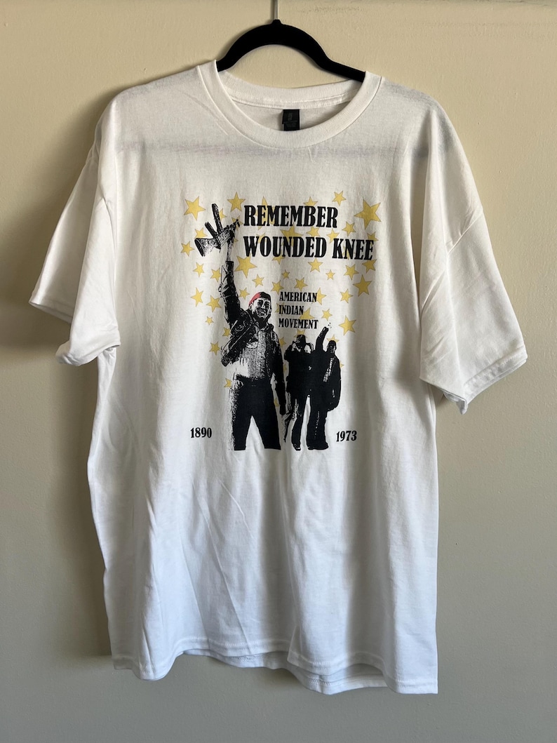Pode incluir: Camiseta branca com uma estampa gr&aacute;fica em preto e branco de uma pessoa segurando um megafone com o texto "Remember Wounded Knee" e "American Indian Movement" com estrelas e os anos 1890 e 1973.