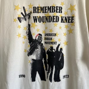Pode incluir: Camiseta branca com uma ilustra&ccedil;&atilde;o em preto e branco de pessoas segurando um rifle. O texto "Remember Wounded Knee" est&aacute; impresso em preto na parte superior da camiseta. O texto "American Indian Movement" est&aacute; impresso em preto abaixo da ilustra&ccedil;&atilde;o. Os anos "1890" e "1973" est&atilde;o impressos em preto na parte inferior da camiseta.
