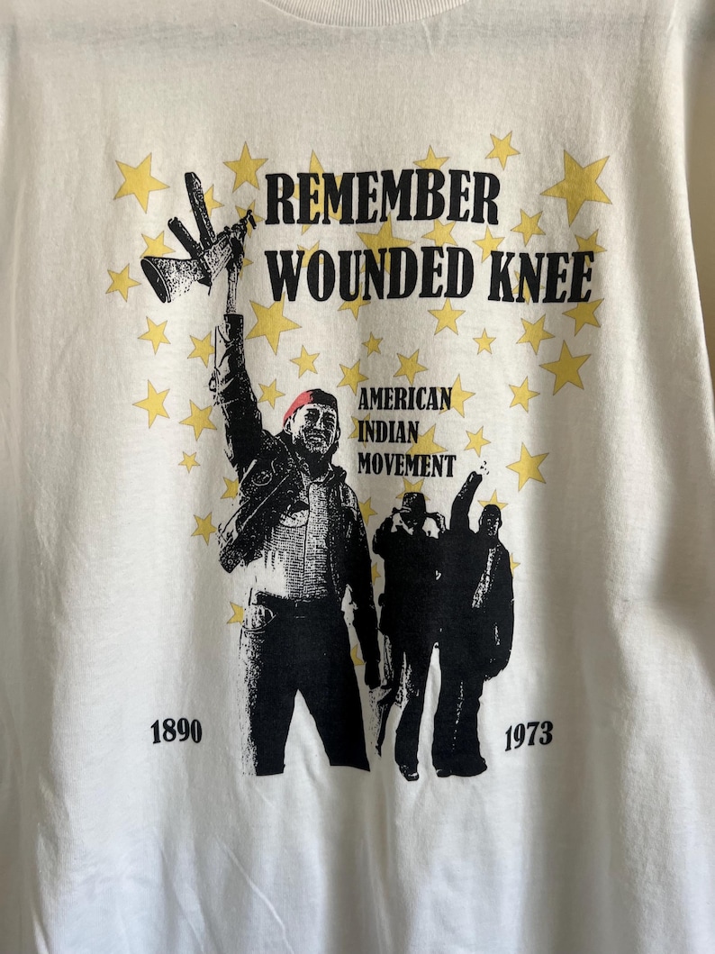 Pode incluir: Camiseta branca com uma ilustra&ccedil;&atilde;o em preto e branco de pessoas segurando um rifle. O texto "Remember Wounded Knee" est&aacute; impresso em preto na parte superior da camiseta. O texto "American Indian Movement" est&aacute; impresso em preto abaixo da ilustra&ccedil;&atilde;o. Os anos "1890" e "1973" est&atilde;o impressos em preto na parte inferior da camiseta.