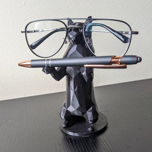 May include: A black, geometric, 3D printed animal figurine holding a pair of black framed glasses and a black pen. The figurine is standing on a black base.
