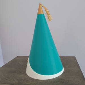 May include: A conical party hat featuring a teal main body with a white curved base. The top of the cone is finished with a metallic gold cap and a matching gold tassel hanging from the peak. The design is smooth and structured in appearance.