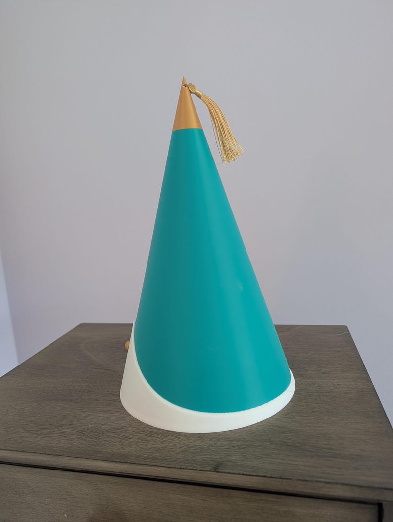 May include: A conical party hat featuring a teal main body with a white curved base. The top of the cone is finished with a metallic gold cap and a matching gold tassel hanging from the peak. The design is smooth and structured in appearance.