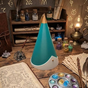 May include: A conical object with a teal body, a gold-colored pointed top, and a white base featuring a small gold stud. The item is positioned on a wooden surface alongside an open book with circular diagrams, a paint palette, and an inkwell.