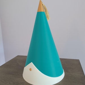 May include: A conical party hat featuring a teal main body, a white base trim with a pointed center, and a gold-colored tip. A small gold stud is centered on the white trim, and a gold tassel is attached to the top of the cone.