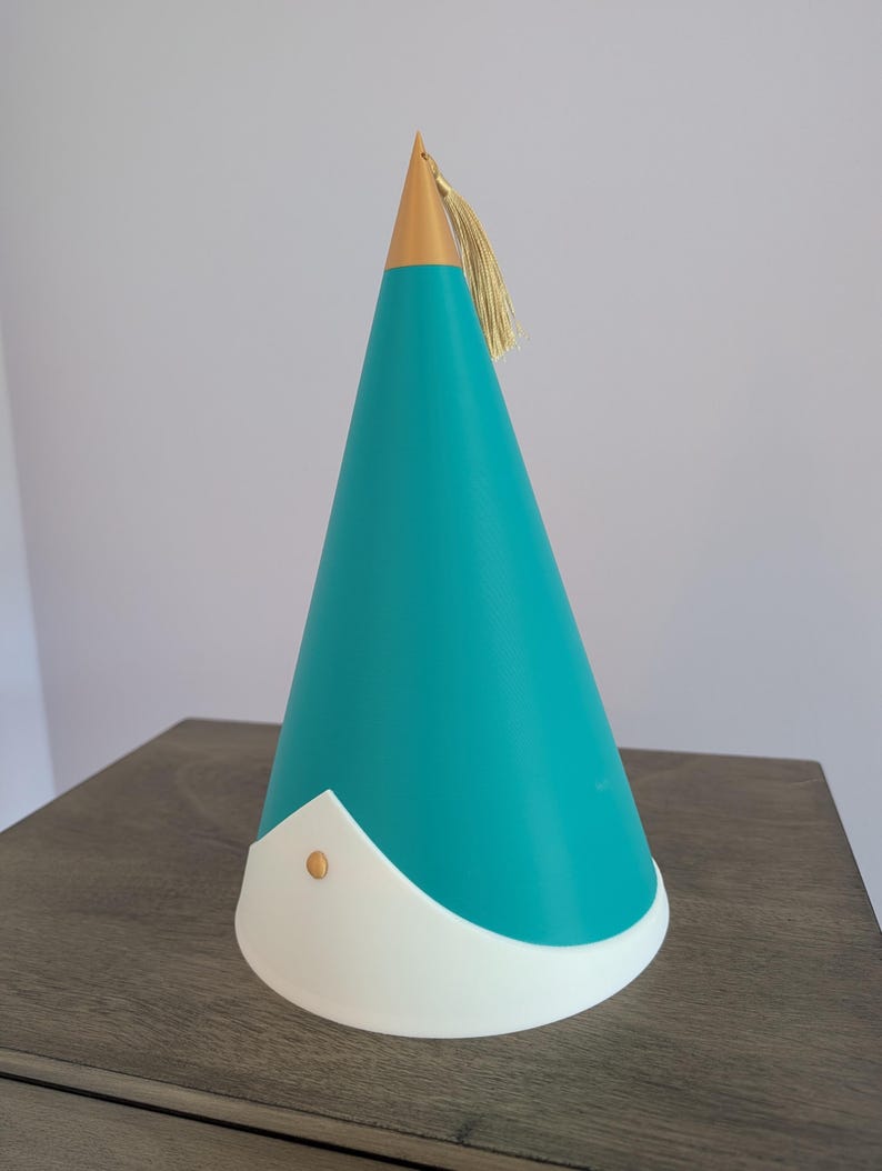 May include: A conical party hat featuring a teal main body, a white base trim with a pointed center, and a gold-colored tip. A small gold stud is centered on the white trim, and a gold tassel is attached to the top of the cone.