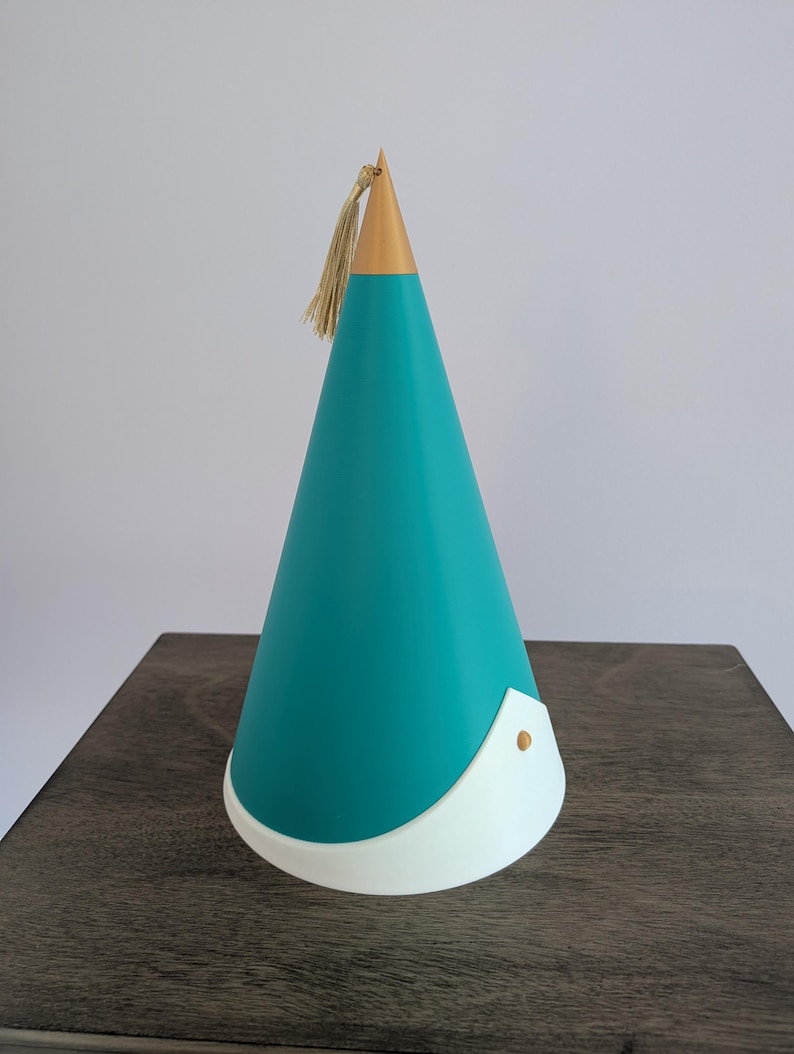 May include: A conical object featuring a teal body, a gold-colored pointed top, and a white base trim. A small gold-colored tassel hangs from the apex of the cone, and a single gold-colored circular stud is attached to the white base section.