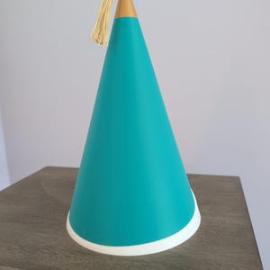 May include: A conical party hat featuring a vibrant teal body, a solid gold-colored pointed top, and a delicate gold tassel attached to the apex. The base of the hat is finished with a clean white circular trim, creating a distinct contrast.