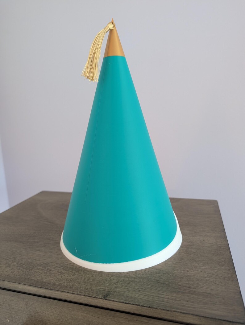 May include: A conical party hat featuring a vibrant teal body, a solid gold-colored pointed top, and a delicate gold tassel attached to the apex. The base of the hat is finished with a clean white circular trim, creating a distinct contrast.