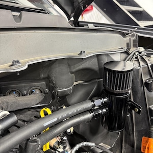 以下が含まれることがあります： 黒いオイルキャッチタンク、黒いフィルターとフィッティング付き、エンジンルームに取り付けられています。画像は、ホースやその他の部品を含むエンジンコンポーネントのクローズアップを示しています。キャッチタンクは、オイルやその他の汚染物質を収集するように設計されています。