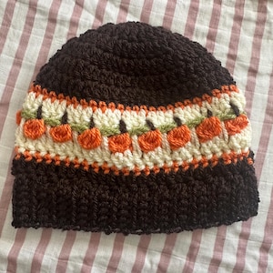 Op de afbeelding: Een donkerbruine gehaakte beanie met een decoratieve band met oranje pompoenen, crème en groene accenten. De hoed heeft een geribbelde textuur en is ideaal voor de herfst. Een warm en modieus accessoire voor koeler weer.