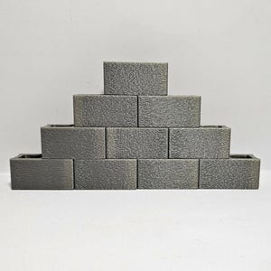 Könnte beinhalten: Eine Pyramide aus grauen, rechteckigen Pflanzgefäßen mit strukturierter Oberfläche. Die Pflanzgefäße sind in einer abgestuften Formation angeordnet, wobei jede Ebene kleiner wird. Der Hintergrund ist eine schlichte weiße Oberfläche.