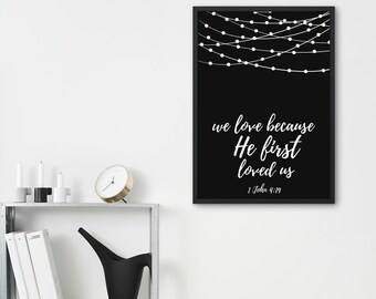 May include: Black framed wall art with white text that reads "we love because He first loved us" and "1 John 4:19". The top of the art features a string of white lights against a black background.