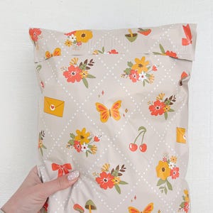 May include: A beige poly mailer with a repeating pattern of white polka dots, orange and yellow flowers, butterflies, cherries, and mushrooms. The mailer has a white seam down the center.