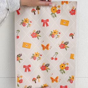 May include: A beige mailing envelope with a repeating pattern of red bows, yellow flowers, green leaves, red cherries, orange butterflies, and brown mushrooms. The envelope has a white dotted background.