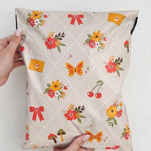 May include: A beige poly mailer with a repeating pattern of red, yellow, and orange flowers, butterflies, cherries, mushrooms, and envelopes with hearts. The mailer has a white polka dot pattern and a diamond pattern.