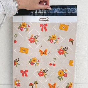 May include: A beige poly mailer with a repeating pattern of flowers, butterflies, cherries, mushrooms, and envelopes. The mailer has a white label with the text "Lucky Duck Packaging" in black.