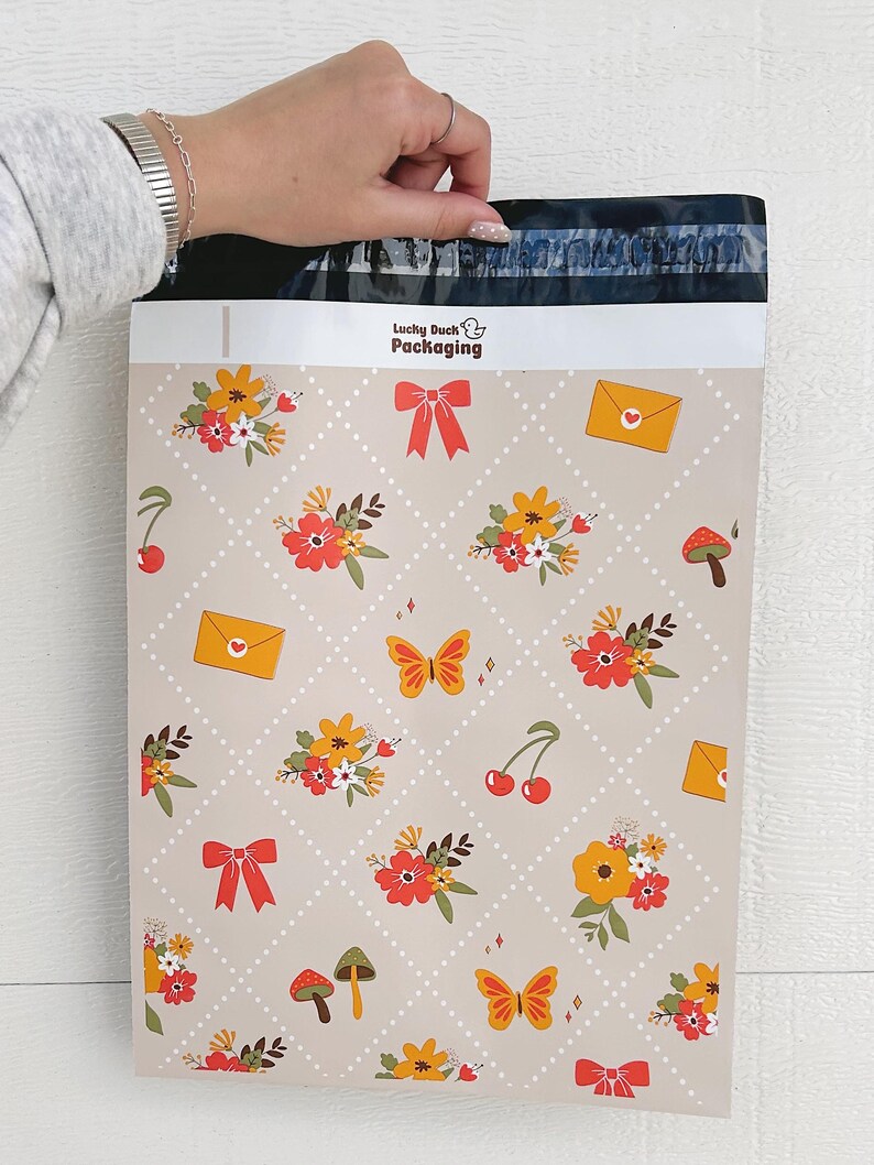May include: A beige poly mailer with a repeating pattern of flowers, butterflies, cherries, mushrooms, and envelopes. The mailer has a white label with the text "Lucky Duck Packaging" in black.