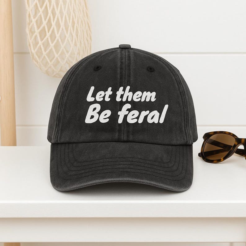 Może przedstawiać: Czarna czapka z daszkiem z białym napisem "Let them Be feral". Czapka ma zakrzywiony daszek i klasyczny sześciopanelowy design. Okulary przeciwsłoneczne po prawej stronie.