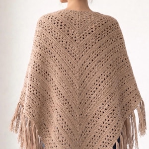 May include: A tan crochet poncho with a fringed hem. The poncho features a V-neck design and a textured pattern with openwork details. The model is wearing the poncho, back view. The poncho is a neutral colour.