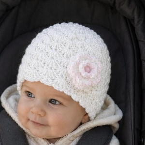 May include: A baby wearing a white crocheted hat with a pink flower accent, seated in a dark gray stroller. The baby is also wearing a cream-colored hooded jacket with a zipper and dark gray safety straps.