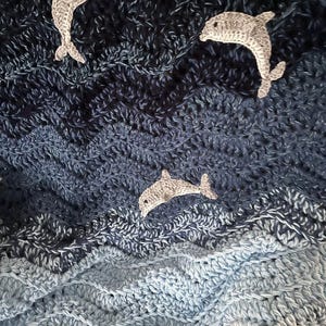 May include: A crocheted blanket in shades of blue, resembling ocean waves. Three gray crocheted dolphins are scattered across the surface. The blanket's texture is visible, with a wavy pattern. The overall theme is nautical.