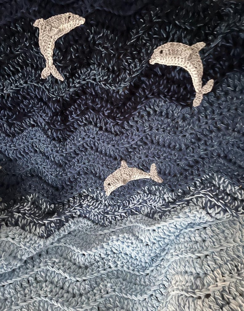 May include: A crocheted blanket in shades of blue, resembling ocean waves. Three gray crocheted dolphins are scattered across the surface. The blanket's texture is visible, with a wavy pattern. The overall theme is nautical.