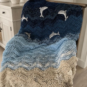 May include: A handmade crochet blanket in shades of blue and tan, depicting an ocean scene. The blanket features a wave pattern with embroidered dolphins and starfish. The blanket is draped over a wooden chair, showcasing its texture and design.