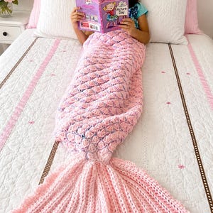 Peut inclure: Une couverture en forme de queue de sirène rose crochetée, avec un motif d'écailles texturées et une longue queue fluide. La couverture est sur un lit avec une couette blanche et des oreillers roses. Un livre intitulé "Missy's Picture Day" est lu.