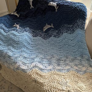 May include: A handmade crochet blanket depicting an ocean scene. The blanket features shades of blue and cream, with wave-like patterns. Dolphins and starfish are appliqued on the blanket, adding a nautical theme. The blanket is draped over a chair.