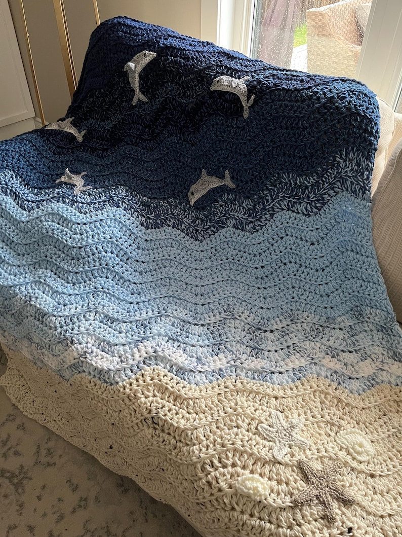 May include: A handmade crochet blanket depicting an ocean scene. The blanket features shades of blue and cream, with wave-like patterns. Dolphins and starfish are appliqued on the blanket, adding a nautical theme. The blanket is draped over a chair.