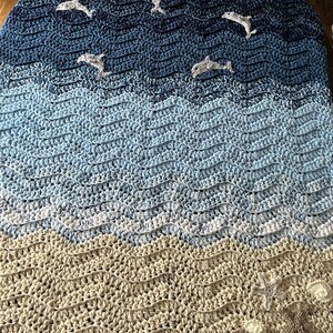 May include: A handmade crochet blanket with a seascape design. The blanket features a gradient of blues and beige, with dolphin appliqu&eacute;s swimming across the waves. The bottom edge has starfish and seashell embellishments.