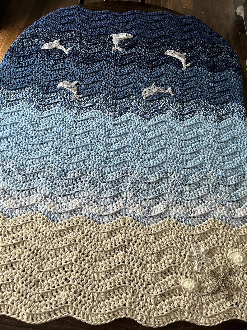 May include: A handmade crochet blanket with a seascape design. The blanket features a gradient of blues and beige, with dolphin appliqu&eacute;s swimming across the waves. The bottom edge has starfish and seashell embellishments.
