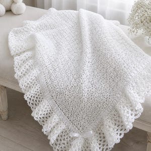 May include: A white crocheted baby blanket with a ruffled edge and a satin ribbon detail. The blanket is folded on a bench next to a white teddy bear and a vase of flowers. The blanket is made of a soft, textured yarn.