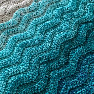 May include: Close-up of a crocheted blanket with a wave-like pattern. The blanket transitions from light gray to shades of blue and teal. The texture of the yarn is visible, creating a cozy and textured appearance. The blanket is suitable for home decor.