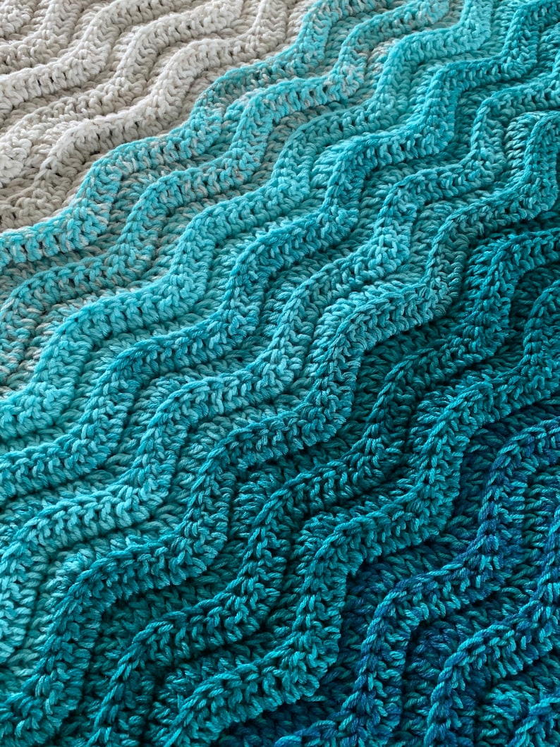 May include: Close-up of a crocheted blanket with a wave-like pattern. The blanket transitions from light gray to shades of blue and teal. The texture of the yarn is visible, creating a cozy and textured appearance. The blanket is suitable for home decor.