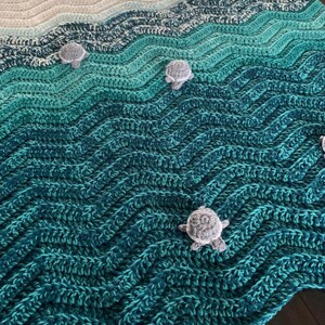 May include: Crocheted blanket in shades of blue and green, resembling ocean waves. Features small, gray crocheted turtle accents scattered across the surface. The blanket's design evokes a coastal or nautical theme.