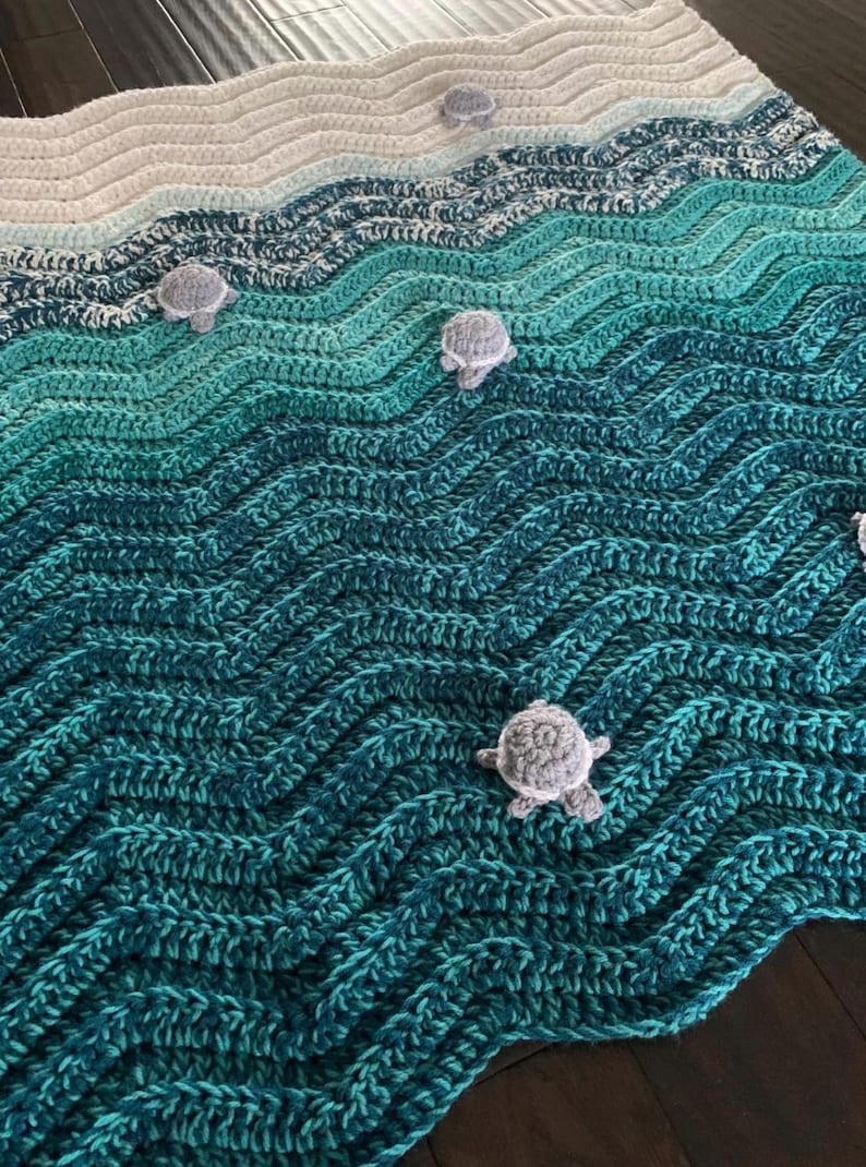 May include: Crocheted blanket in shades of blue and green, resembling ocean waves. Features small, gray crocheted turtle accents scattered across the surface. The blanket's design evokes a coastal or nautical theme.
