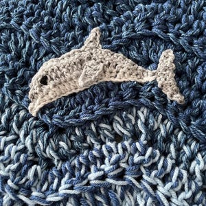 May include: A crocheted blanket with a dolphin applique. The blanket is made of blue yarn in various shades, creating a textured, ocean-like appearance. The dolphin is a light gray color with a black eye.