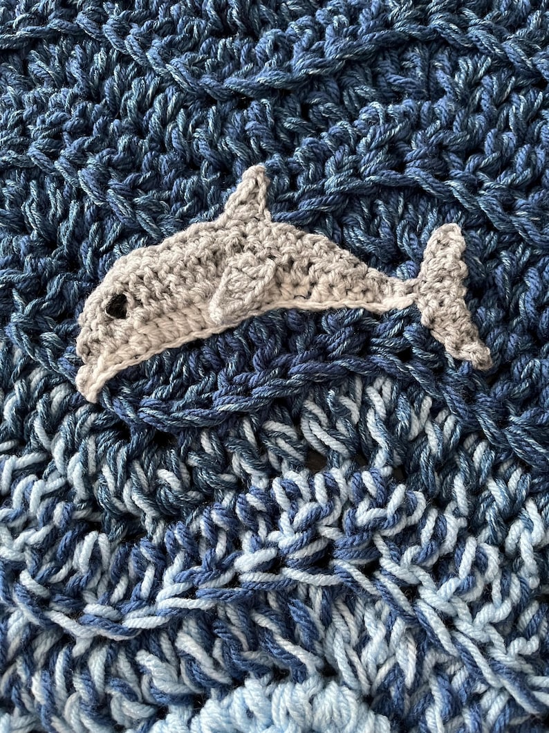 May include: A crocheted blanket with a dolphin applique. The blanket is made of blue yarn in various shades, creating a textured, ocean-like appearance. The dolphin is a light gray color with a black eye.