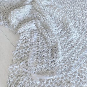 May include: A white, crocheted blanket with a delicate open weave. The blanket features a scalloped edge and is adorned with a white satin ribbon tied in a bow. The soft, textured blanket is ideal for a baby or as a decorative throw.